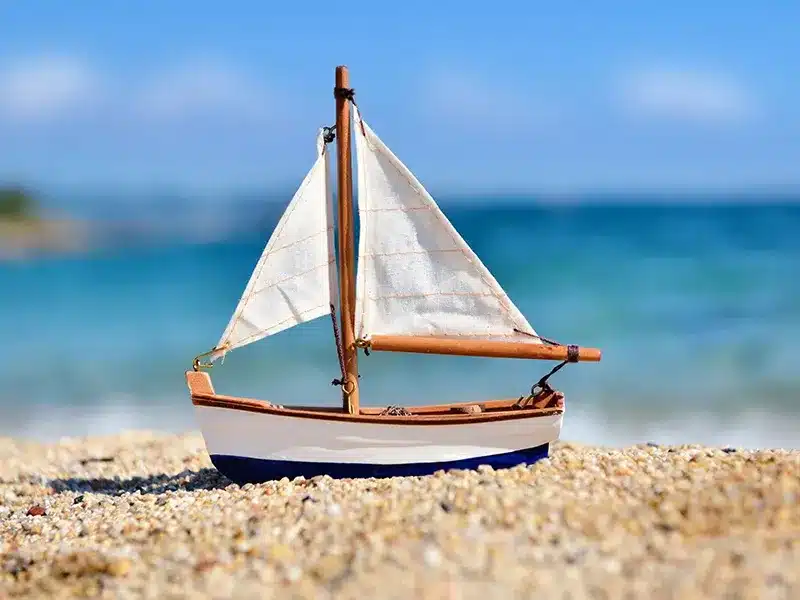 Model sailboat on sandy beach with ocean backdrop.
